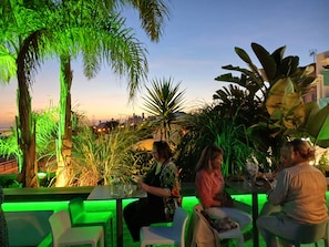An outdoor seating area with tables and chairs, surrounded by greenery and a cityscape in the background.