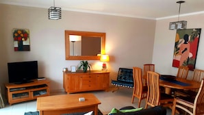 Uma sala de estar com televisão, mobiliário de madeira, uma mesa de jantar e um espelho na parede.