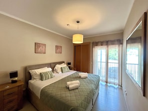 A bedroom with a bed, bedside tables, a hanging light fixture, and a view through a window.