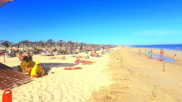 Uma praia com espreguiçadeiras, guarda-sóis, um posto de salva-vidas e uma faixa de areia.