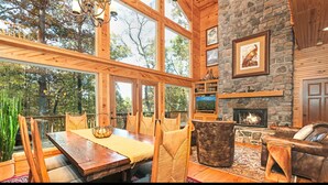 A spacious room with a large wooden dining table, comfortable seating, a stone fireplace, and large windows offering a view of the surrounding trees. - MOUNTAIN HOME--GOLD STAR WINNER!--A peaceful place to breathe in, breathe out... (Hot Springs Village)
