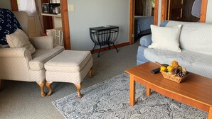 Una sala de estar acogedora con sofá, sillón y reposapiés, una mesita con cesta de frutas y una alfombra estampada.