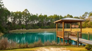Un porche de madera con techo metálico que da a un lago, rodeado de árboles y zonas verdes.