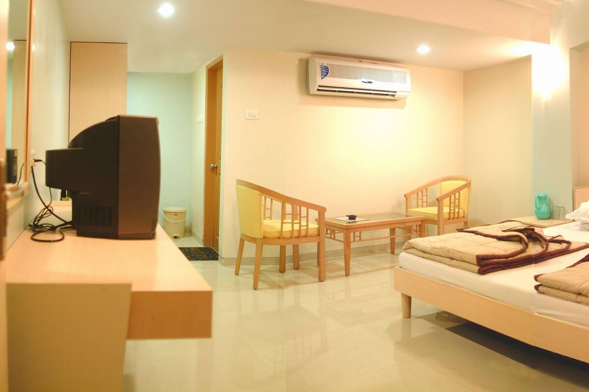 A hotel room with a bed, a desk, two chairs, a television, and an air conditioning unit.
