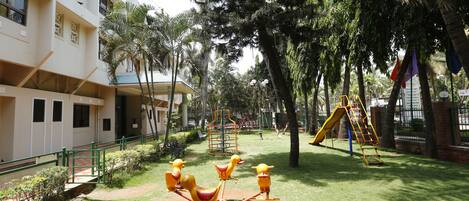 Un parque infantil con columpios y toboganes, rodeado de árboles y un edificio de fachada blanca.