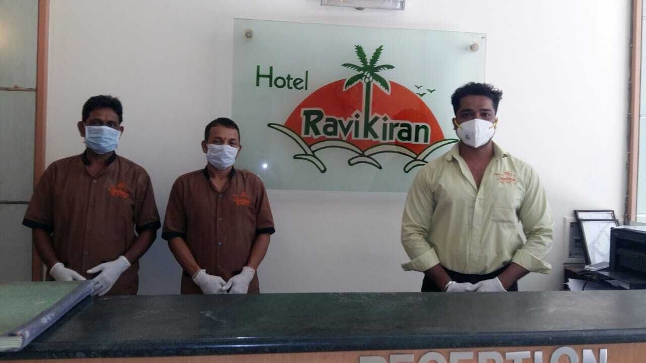 Three individuals standing at a reception desk with a sign for Hotel Ravikiran in the background.