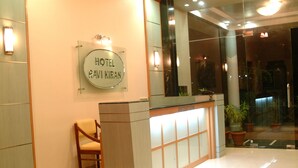 Zona de recepción de un hotel, con mostrador de recepción, una silla de madera y un letrero que dice "HOTEL RAWI KIRAN".