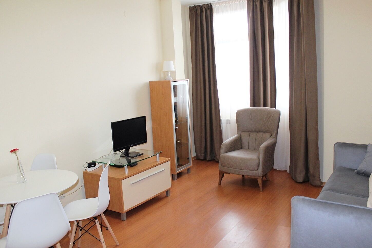 A modern living room with a round glass table, a grey armchair, a grey sofa, and a TV on a wooden cabinet.