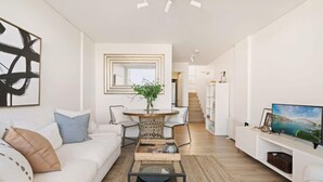 A modern living room with a white sofa, a coffee table, and a TV on a white stand. There is a dining area with a round table and chairs.