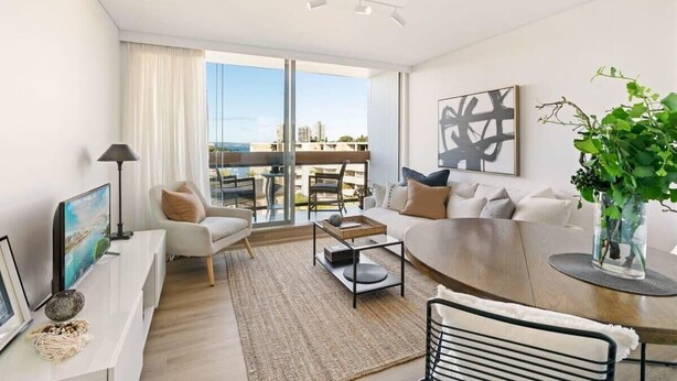 A modern living room with a sofa, coffee table, and a dining area. There is a large window providing a view of the cityscape.