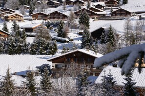Un chalet enneigé, composé de plusieurs bâtiments en bois, entouré d’arbres chargés de neige et situé à proximité d’une piste de ski.
