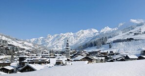 Un village enneigé, avec ses bâtiments traditionnels en bois, un imposant beffroi et, en arrière-plan, des montagnes aux sommets couverts de neige.