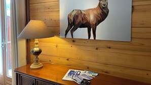 A wooden wall with a framed polygonal deer painting, a lamp, and a table with magazines. - Large, cosy apartment in exceptional location near village. (La Clusaz)