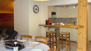 A dining area with a round table set for four, wooden chairs, a clock on the wall, and a kitchen in the background. - Large, cosy apartment in exceptional location near village. (La Clusaz)