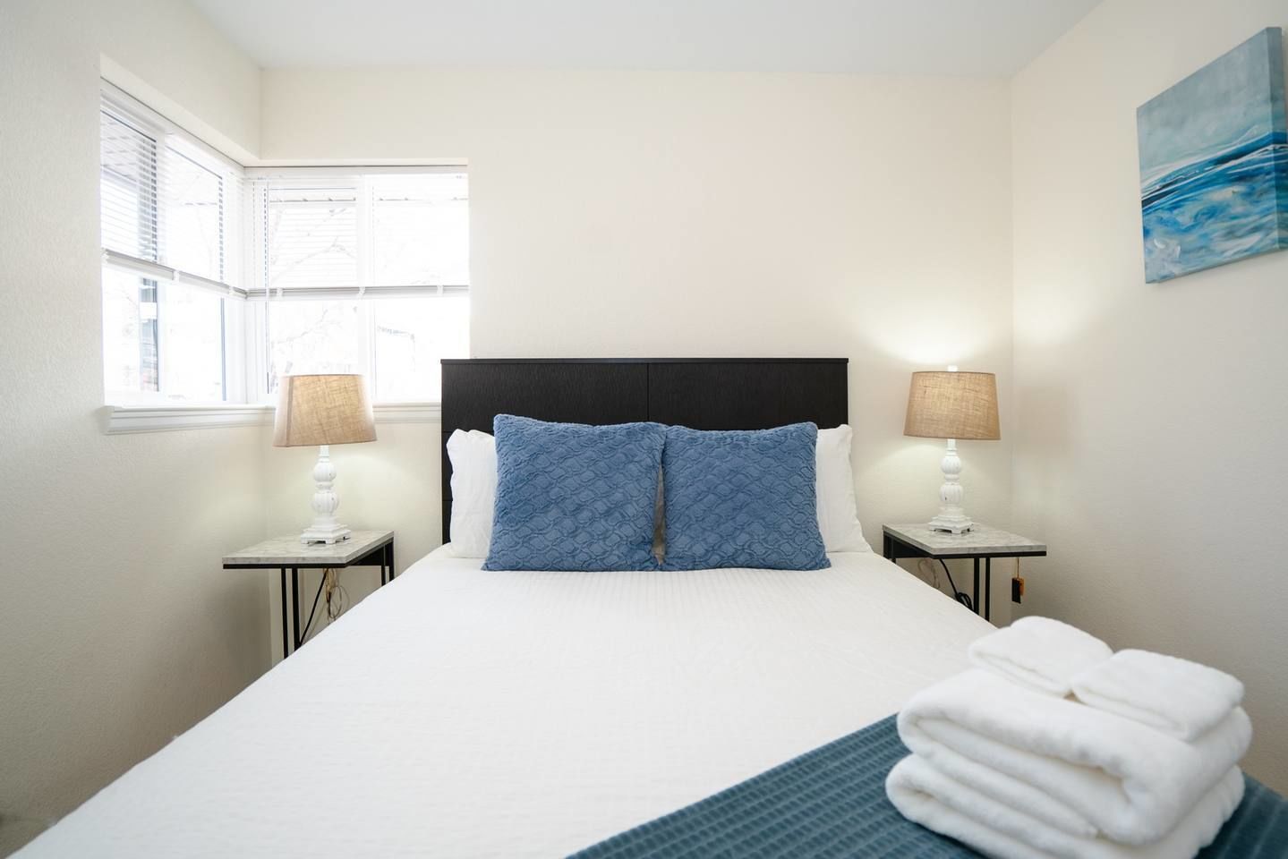 A hotel room with a large bed, two bedside lamps, a window with blinds, and a painting on the wall.