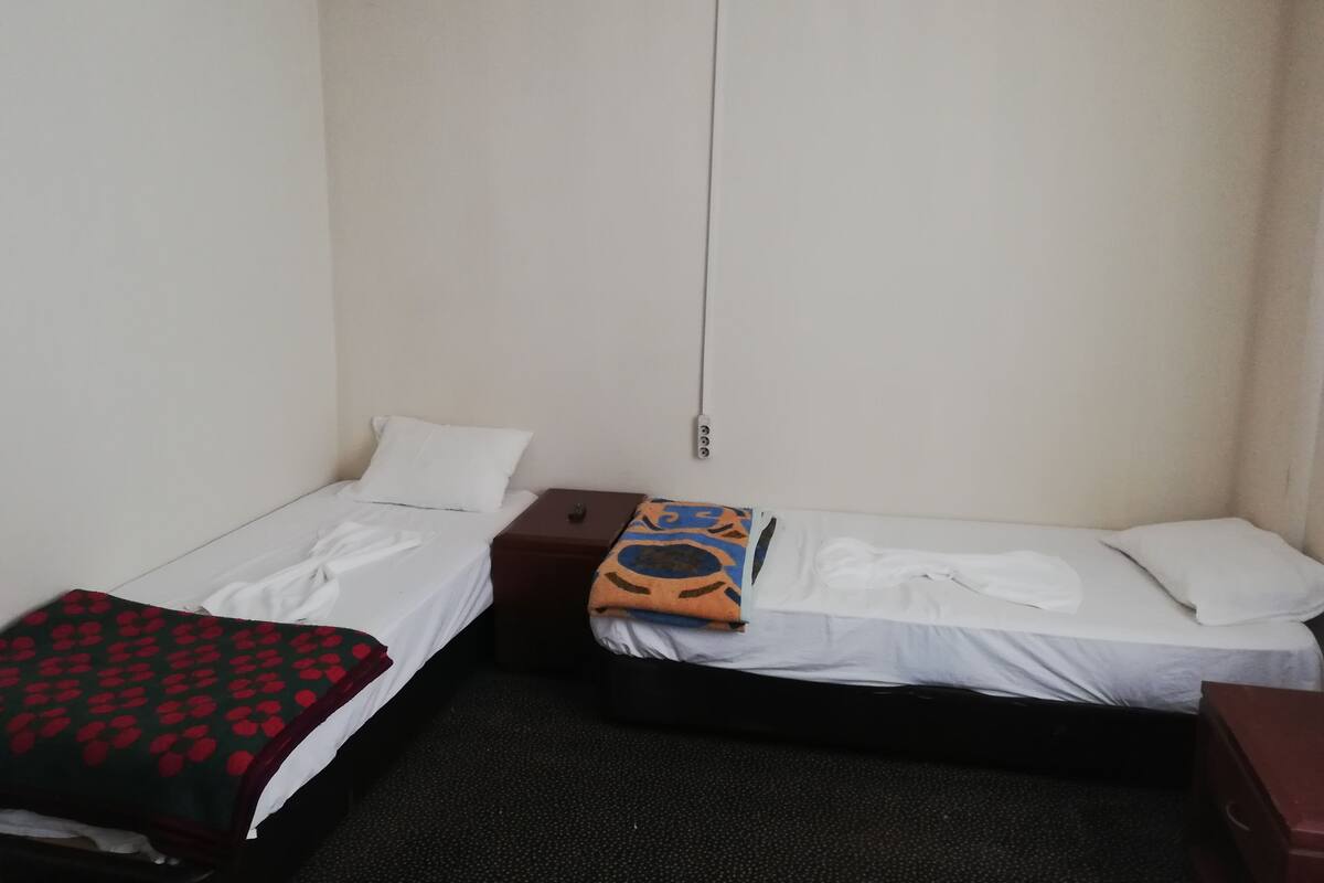 Une petite chambre d’hôtel composée d’une seule pièce, avec deux lits simples, une petite table de chevet en bois et une armoire.