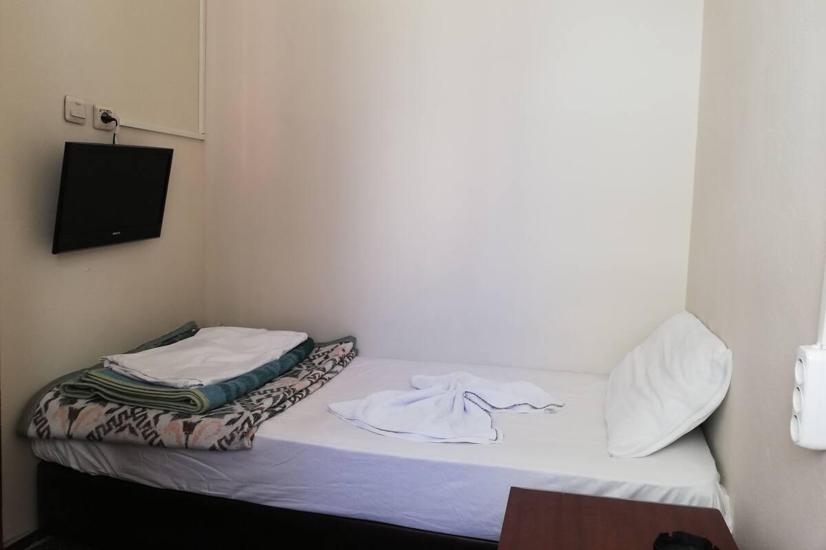 Une petite chambre avec un lit simple, un téléviseur fixé au mur et une table de chevet.