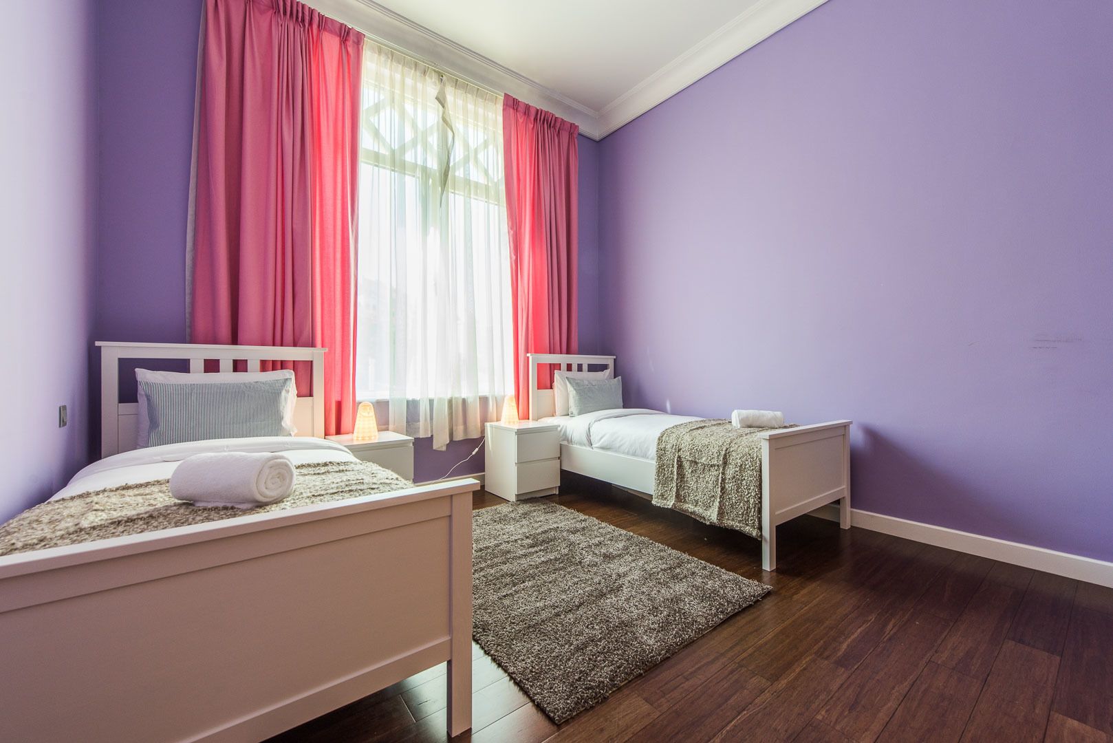 A bedroom with two beds, a window with sheer curtains, and a wooden floor.