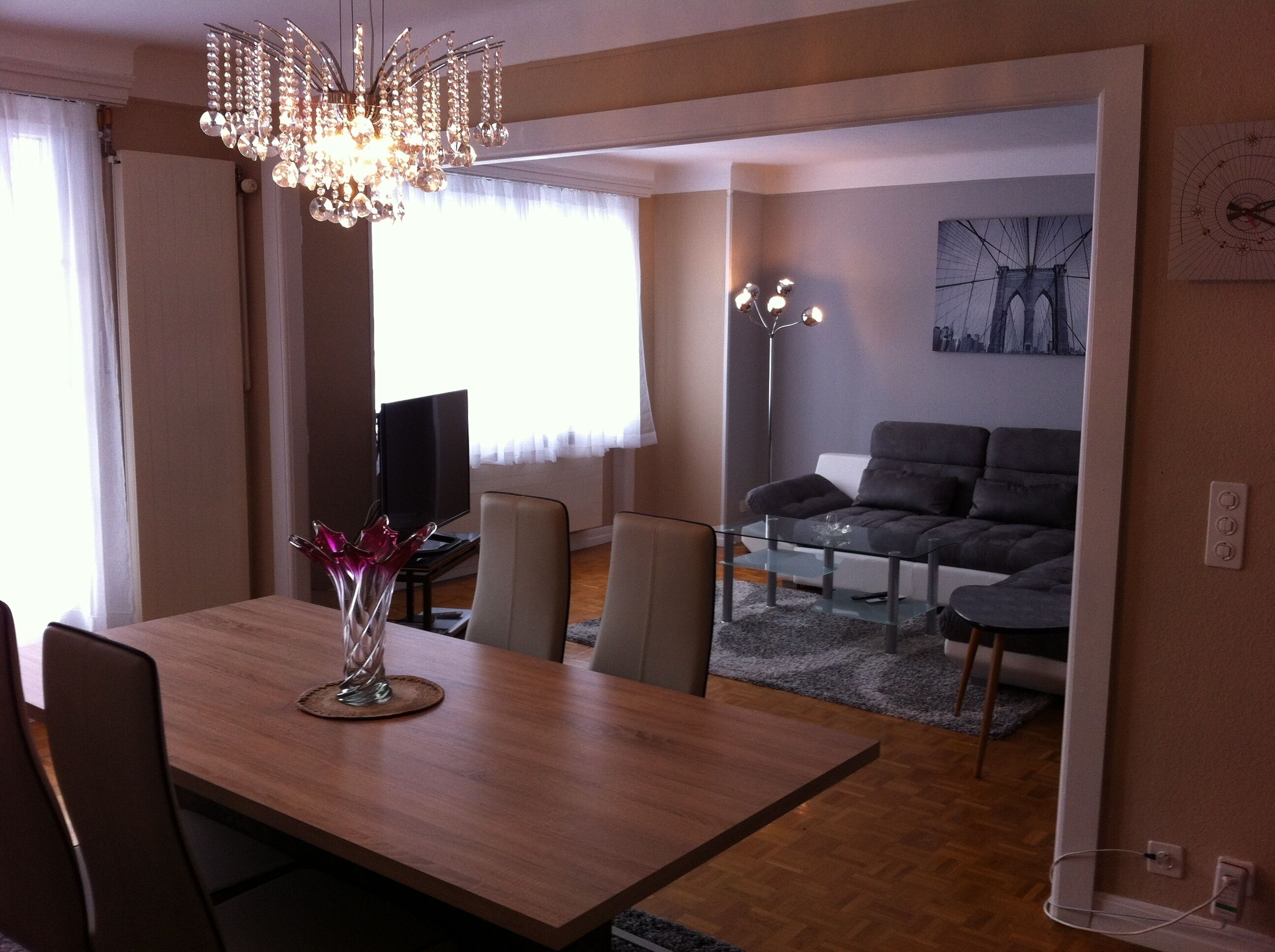 A dining area with a wooden table, chairs, a vase with flowers, a chandelier, and a television in the background.