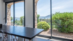 Ein modernes Zimmer mit großem Tisch und Stühlen, großen Fenstern mit Steinbögen und Blick auf Grünflächen sowie ein Gewässer.