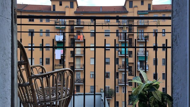 A balcony with a wicker chair and a potted plant, overlooking a multi-story building with balconies and laundry hanging out to dry.