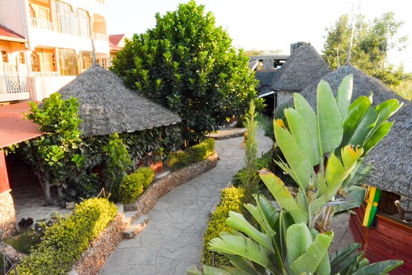 A courtyard with thatched-roof structures, surrounded by lush greenery and a stone pathway.