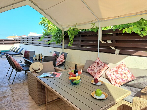 An outdoor patio with wicker furniture, a table with a book, a bowl of fruit, and a bottle of wine.