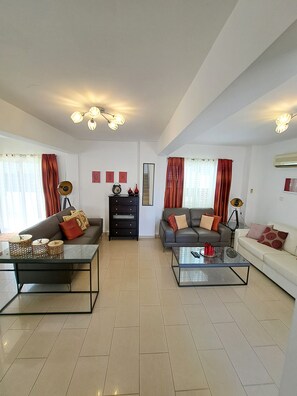 A spacious living room with two sofas, a coffee table, and a TV. There are curtains, a ceiling fan, and wall decorations.