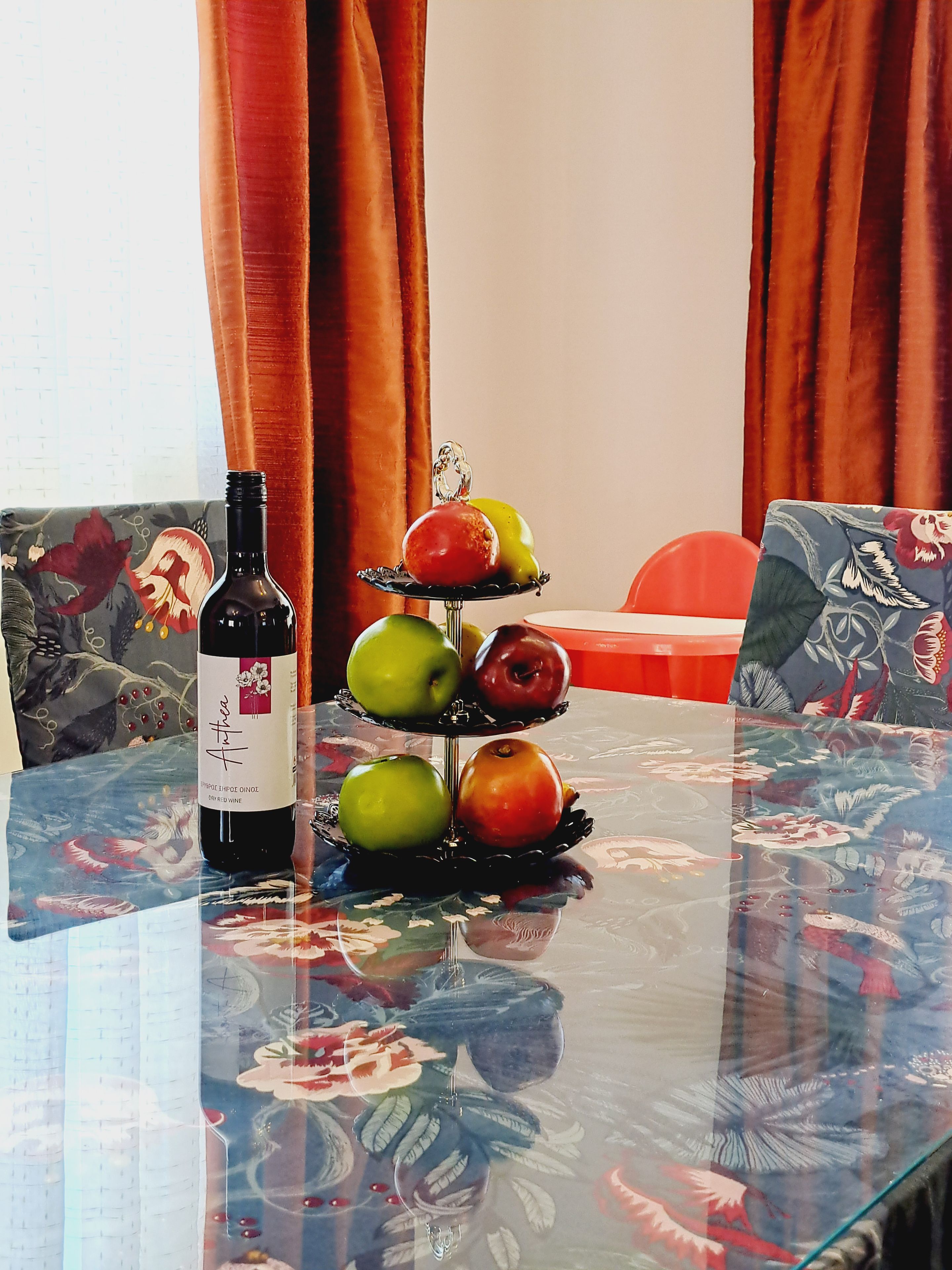 A table with a bottle of wine, a fruit stand, and a floral tablecloth.