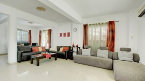 A spacious living room with a black leather sofa, a grey sofa, and a coffee table. There are two windows with curtains, a clock on the wall, and a small table with decorative items. - Villa Anasta near the sea  (Protaras)