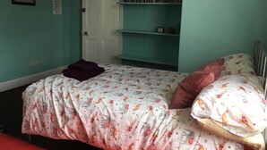 A bedroom with teal walls, a white door, a bed with a floral bedspread, a purple blanket, and a pink pillow. There are shelves with books and a framed picture on the wall. - The Mother Ruin (Bristol)