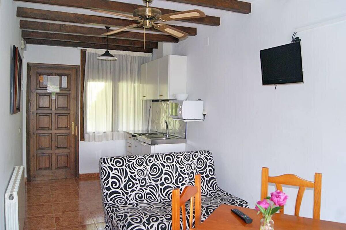 A room with a ceiling fan, a sofa with a black and white pattern, a wooden dining table, and a TV mounted on the wall.