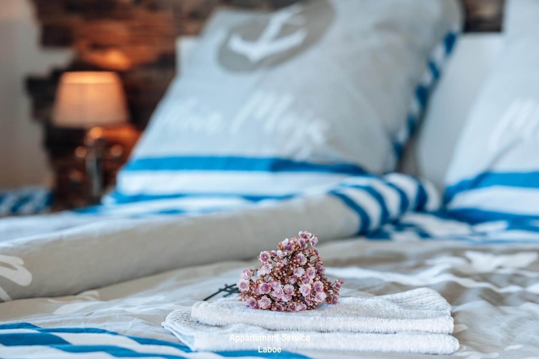 Ein ordentlich bezogenes Bett mit blau-weiß gestreifter Bettwäsche, ein weißes Kopfkissen mit einem weißen Pfeil-Design und ein kleiner Blumenstrauß auf einem gefalteten Handtuch.