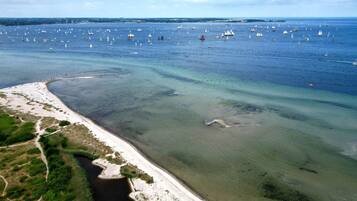 Eine Luftaufnahme einer Küstenlandschaft mit einem Sandstrand, einer kleinen Bucht und zahlreichen Segelbooten auf dem Wasser.