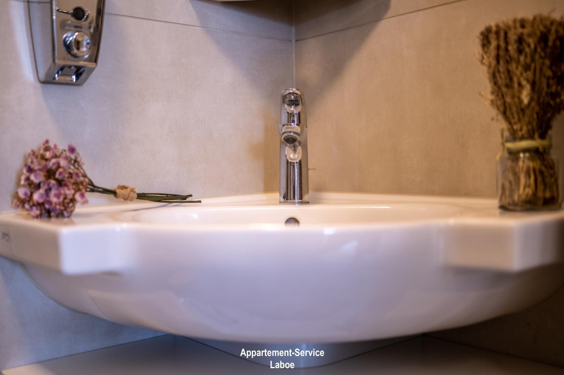 Un lavabo amb un mànec, un vas petit amb flors seques i una lluminària penjada a la paret.