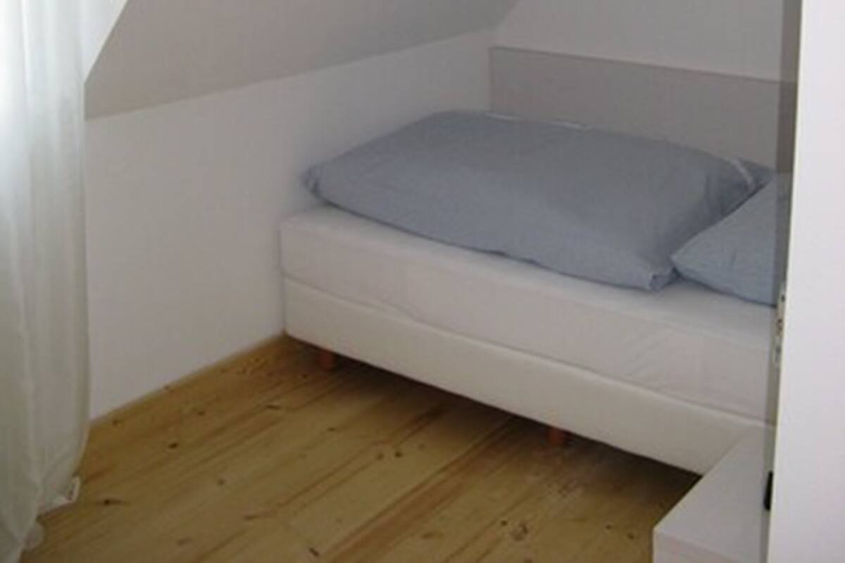 Una habitación pequeña con cama individual, techo inclinado y piso de madera.