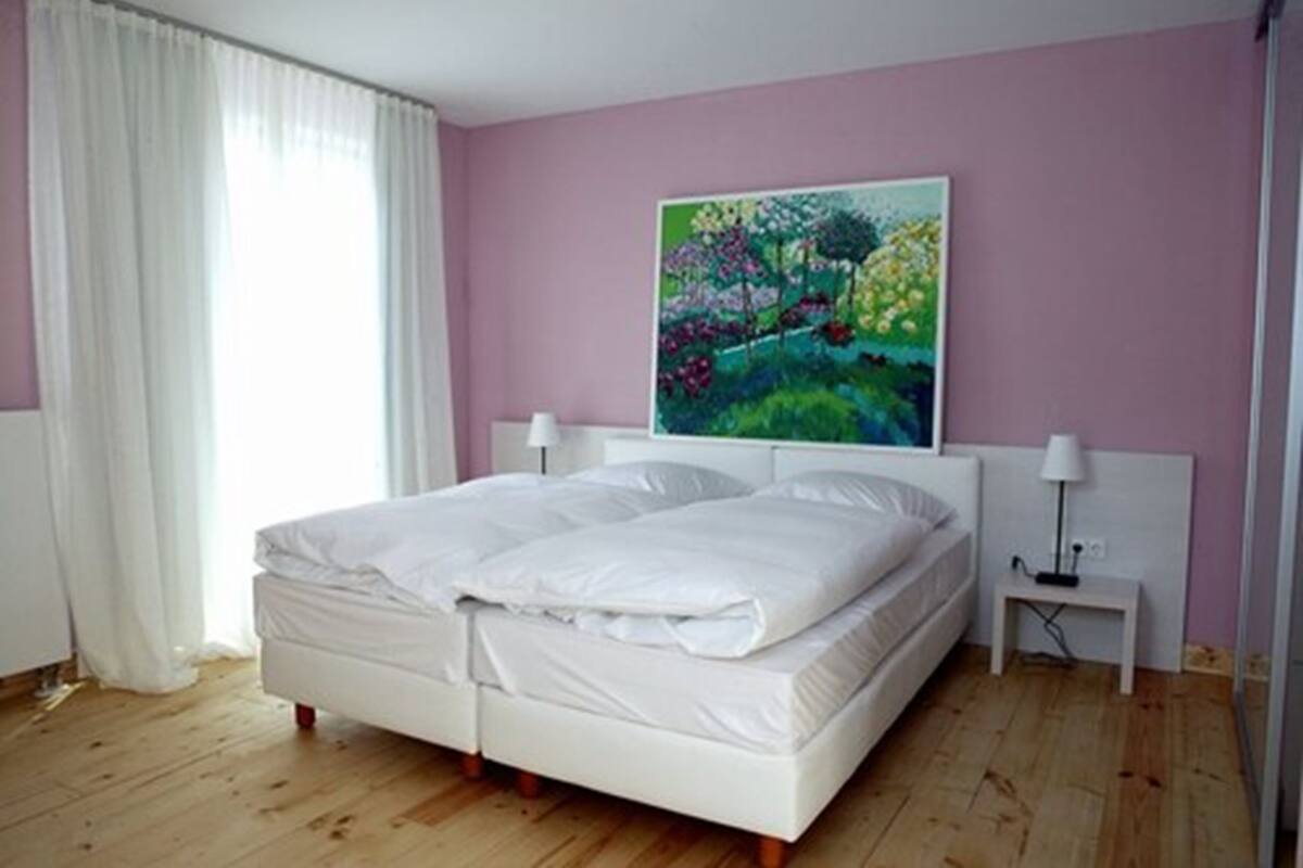 Un dormitorio con cama, mesita de noche, un cuadro y una ventana con cortinas.