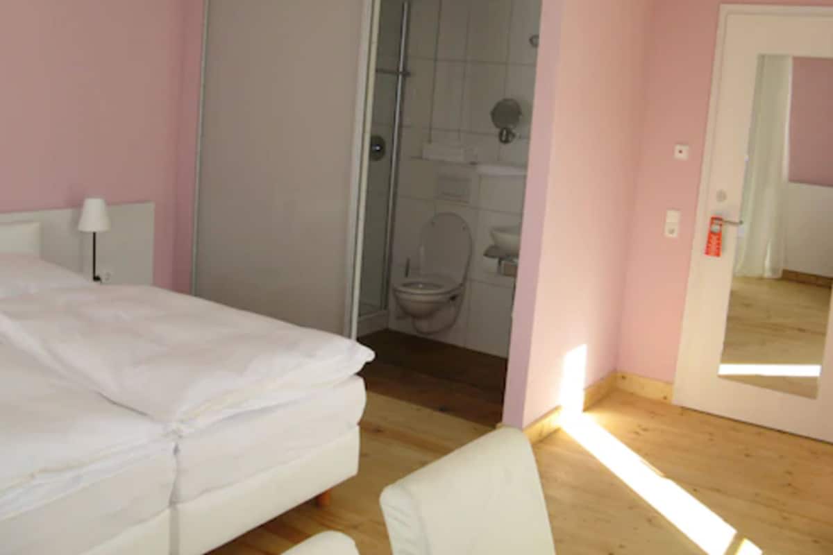 Un dormitorio con cama, una silla, una mesita de noche y un baño visible a través de una puerta abierta.