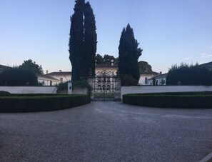 Een statig entree met een ijzeren poort, hoge cipressen aan weerszijden van de poort, en een verzorgde haag op de voorgrond.