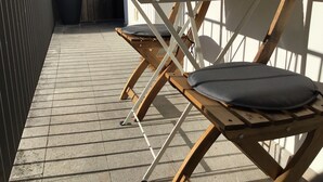 Une table en bois avec un coussin, un vase vert et une plante en pot, sur un balcon dont le sol est recouvert de carreaux.