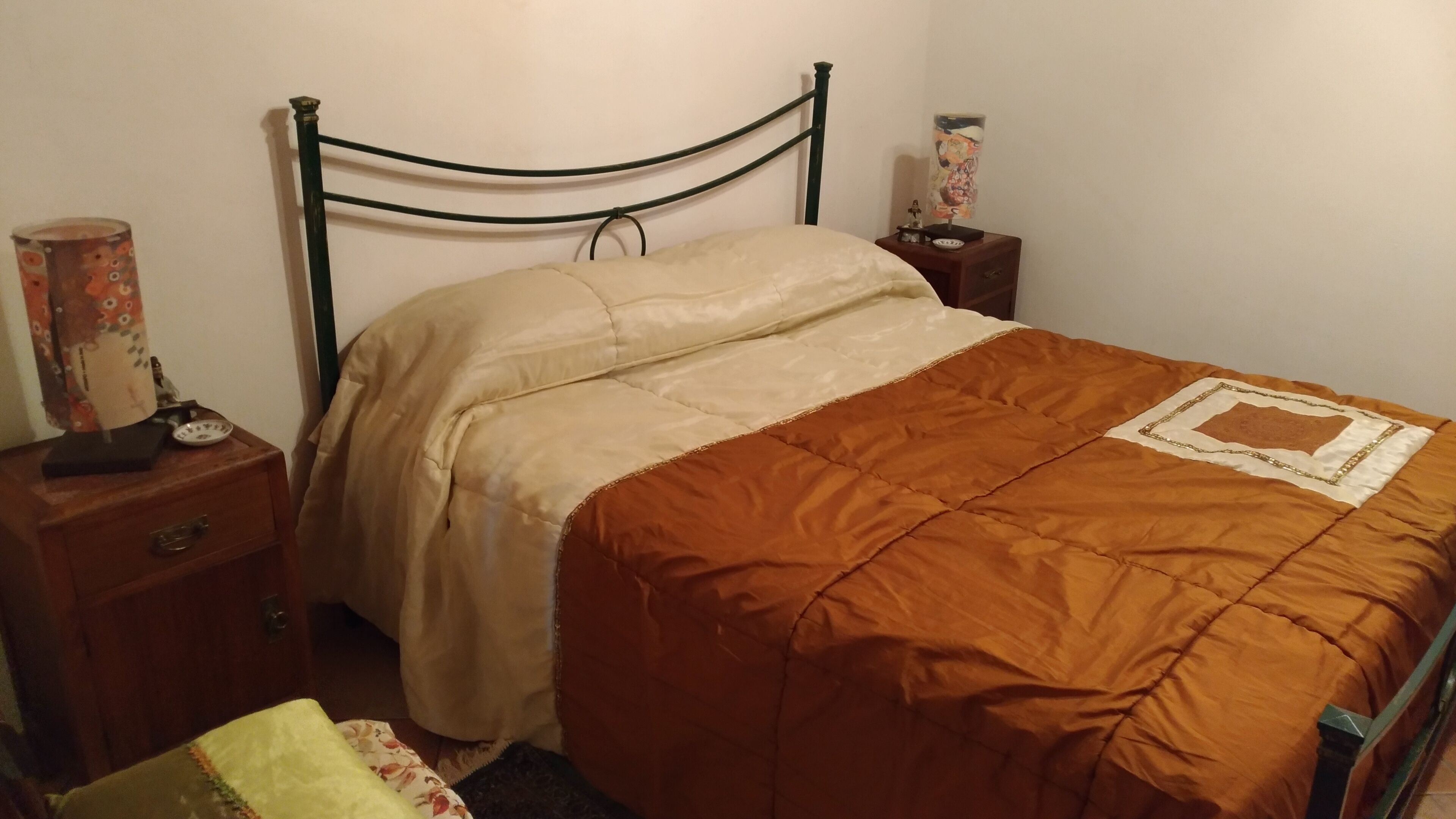 Un letto singolo con struttura in legno, coperta beige e marrone, un comodino con una lampada e un tavolino con un oggetto decorativo.