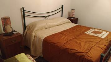 Un letto singolo con struttura in legno, coperta beige e marrone, un comodino con una lampada e un tavolino con un oggetto decorativo.