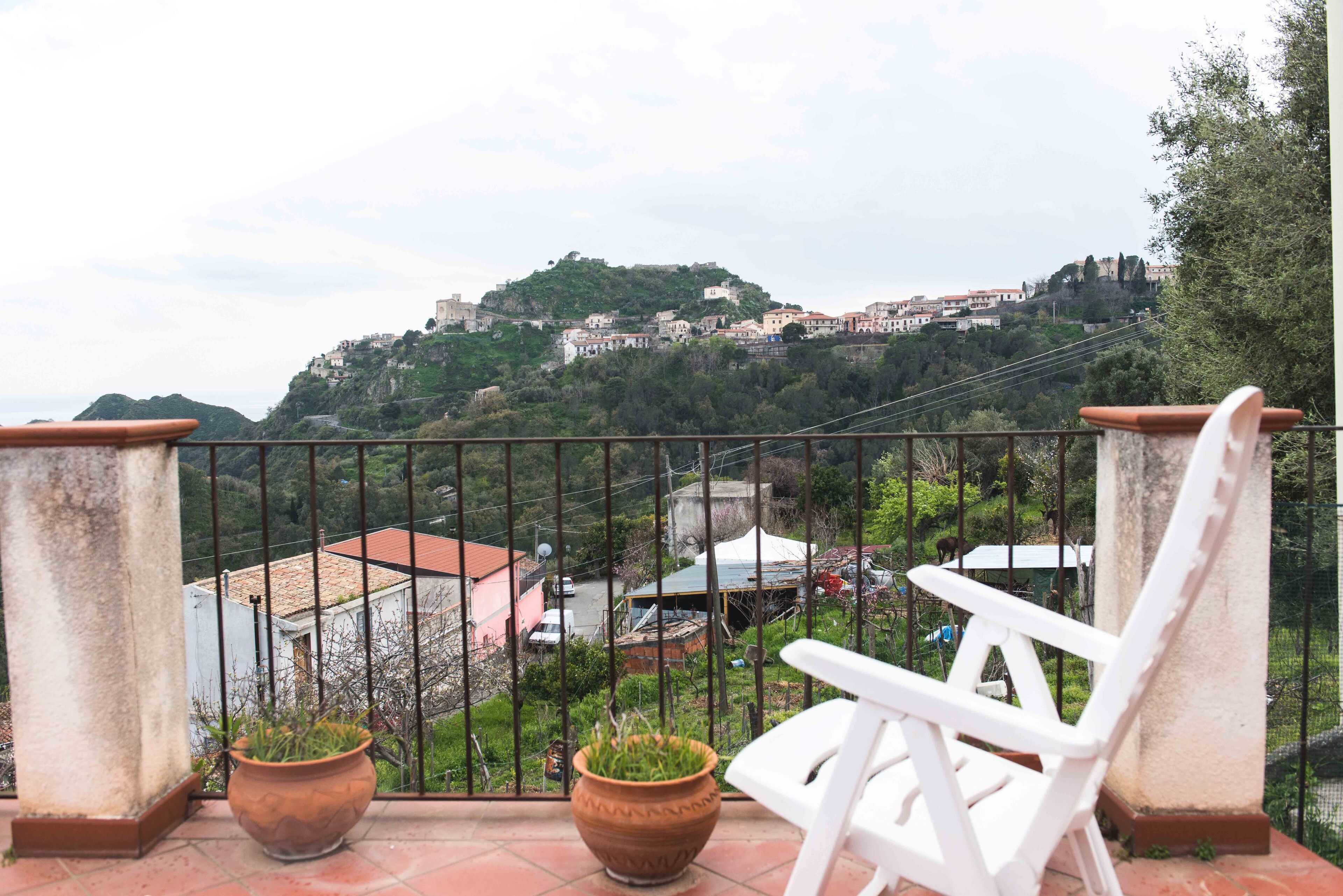 En balkon med en hvid stol og to terracotta-potter, med udsigt over en bakke med bygninger og grøn vegetation.