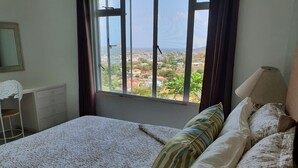 Un dormitorio con cama, una silla, un escritorio y una ventana con vista a un paisaje urbano.