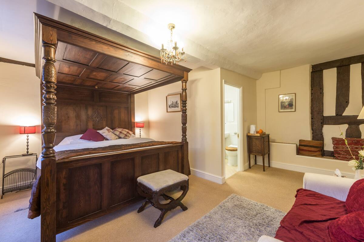 A four-poster bed with a wooden headboard, a small stool, a red upholstered chair, and a white bedside table with a lamp.