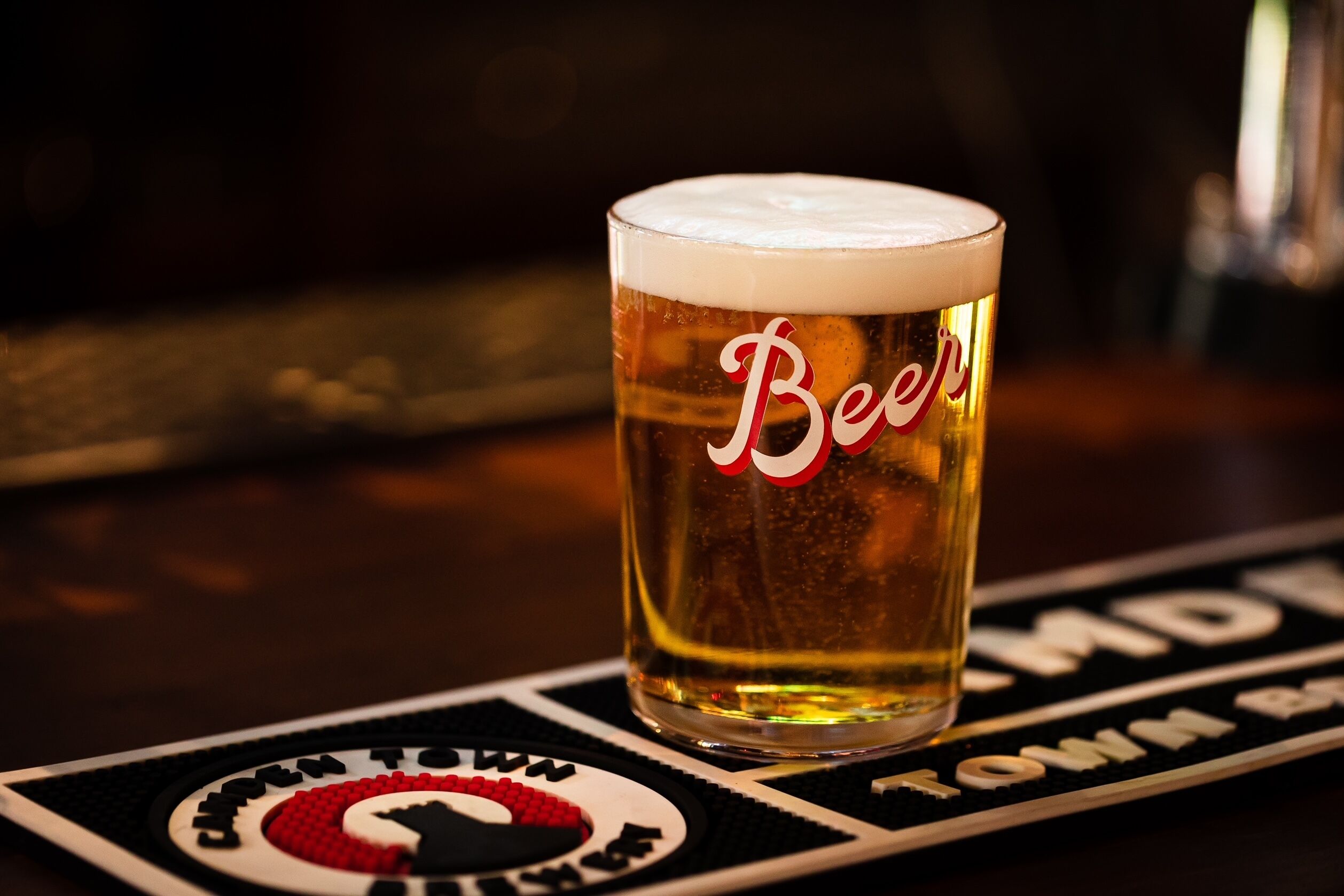 A glass of beer with "Beer" written on it, placed on a coaster with a logo and text.
