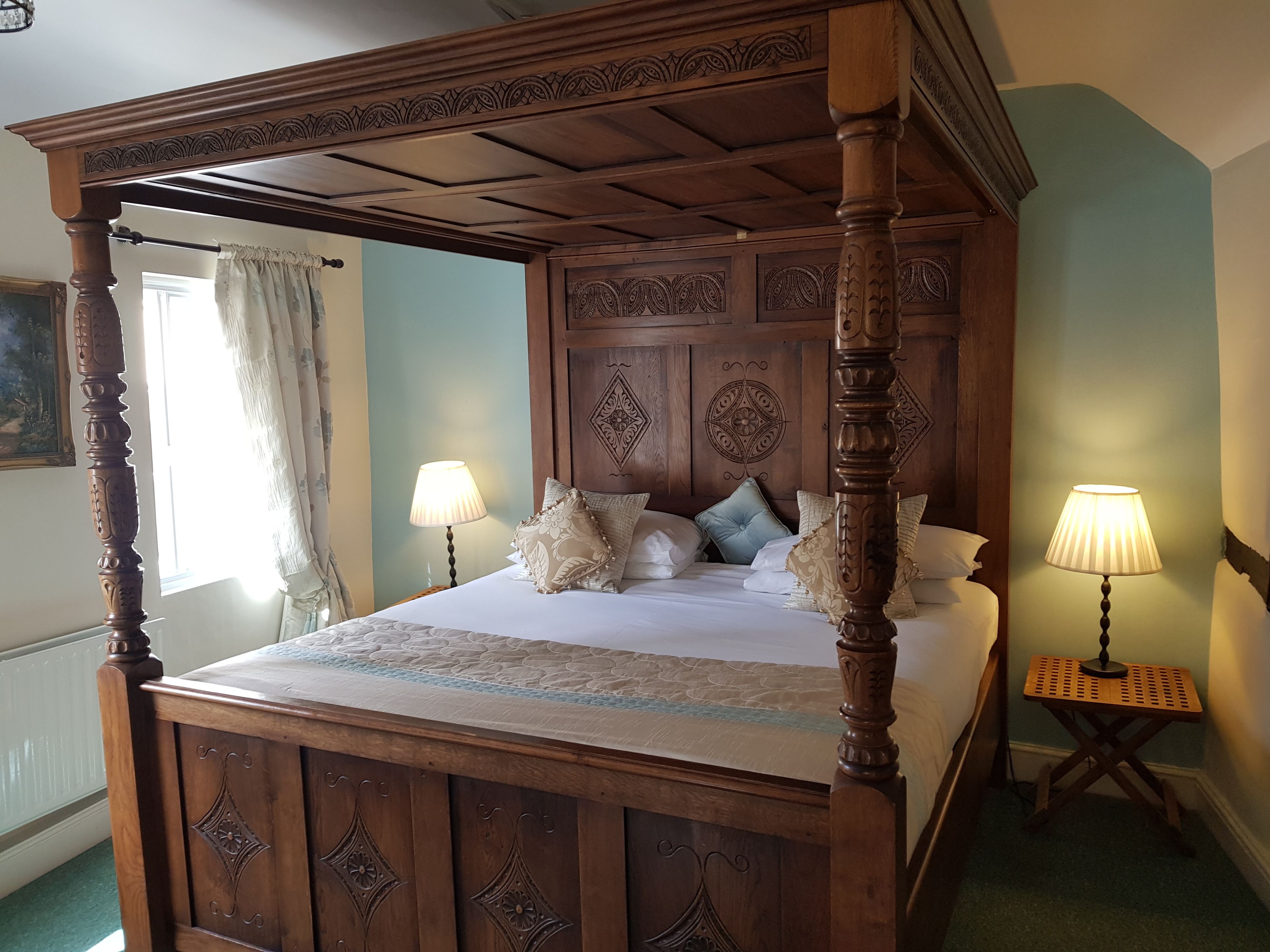 A four-poster bed with intricate woodwork, a nightstand with a lamp, and a painting on the wall.