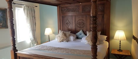 Una cama con dosel y elaborados trabajos en madera, una mesita de noche con lámpara y un cuadro en la pared.