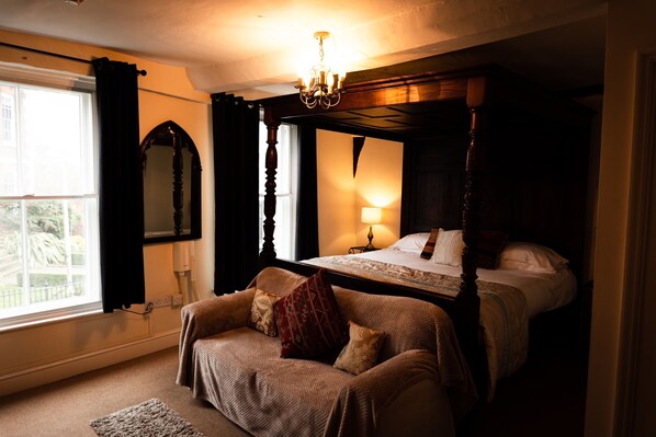 A bedroom with a four-poster bed, a sitting area with a chair and cushions, a bedside table with a lamp, and a large window with curtains.