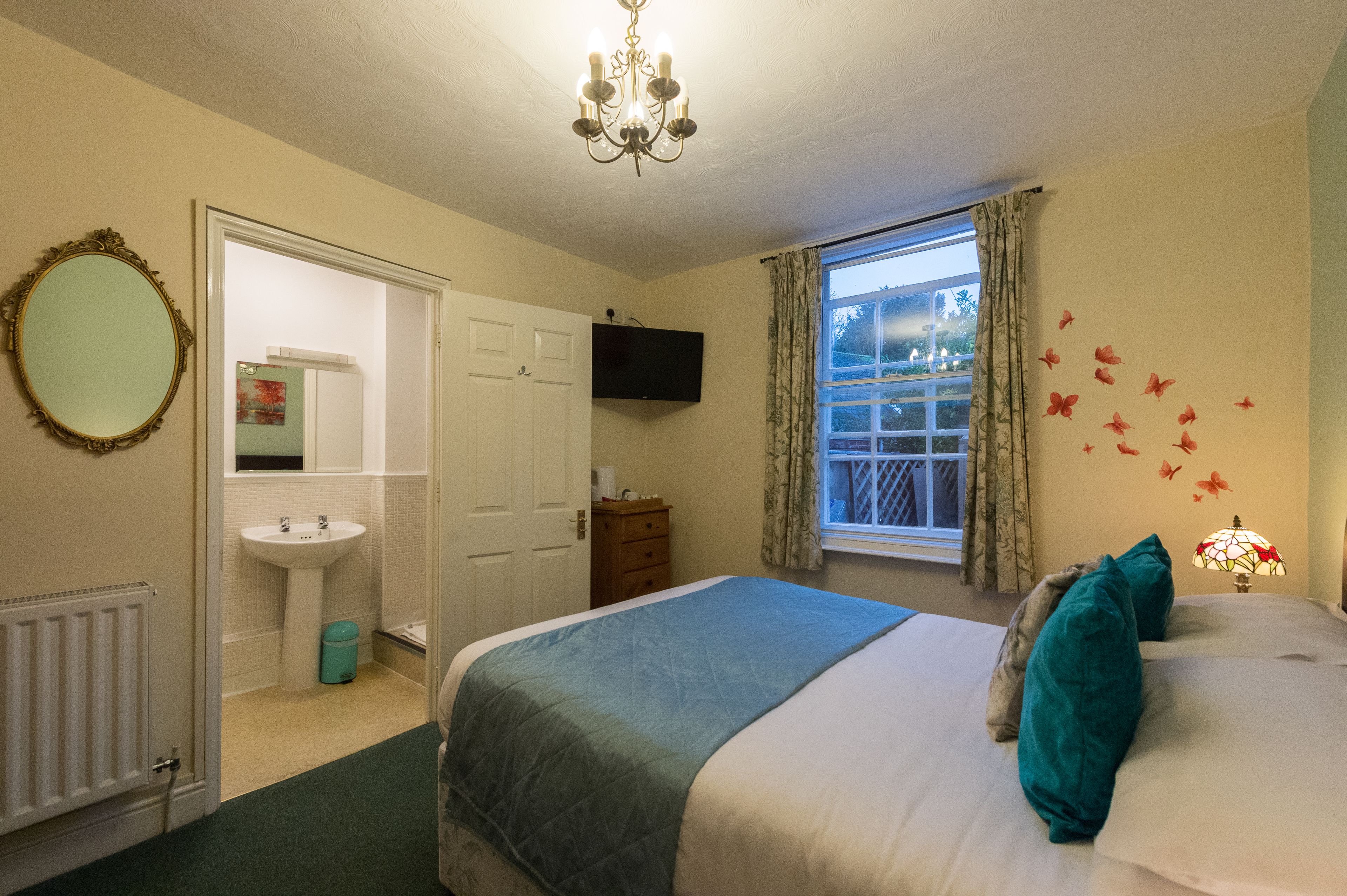 A bedroom with a bed, a bedside table, a lamp, a window with curtains, a television, a mirror, and a bathroom with a sink and toilet.